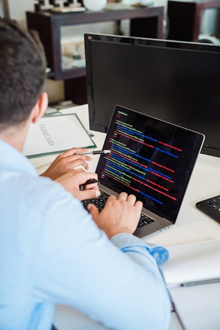 A person wearing a light blue shirt is seated at a desk, working on a laptop displaying lines of colorful code, with an external monitor and various office materials nearby. The workspace includes a k