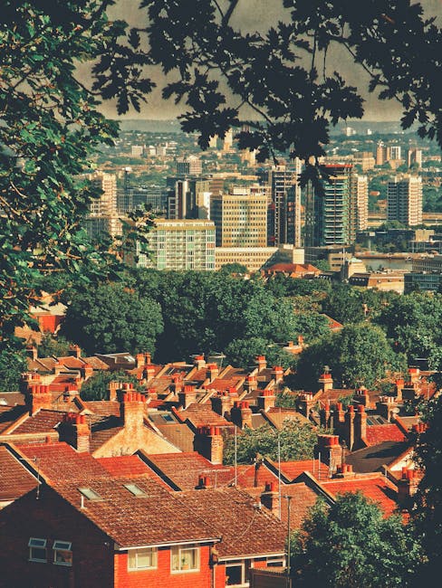 A view of a densely populated urban area with red-brick residential houses in the foreground, featuring tiled roofs and chimneys. Behind the houses, there is a green canopy of trees and a cityscape co