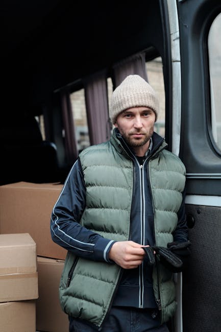 A man wearing a light grey knit beanie, a grey puffy vest over a navy blue jacket, standing inside a moving van with an open door. The van's interior is visible with black seats, and several cardboard