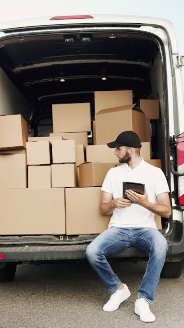 A man wearing a light grey knit beanie, a grey puffy vest over a navy blue jacket, standing inside a moving van with an open door. The van's interior is visible with black seats, and several cardboard